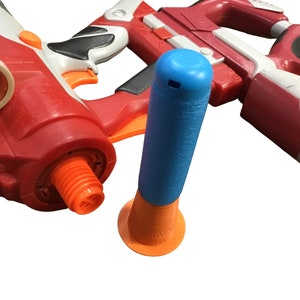 May include: A close-up of a blue and orange toy dart gun with a red and white toy gun in the background. The blue and orange toy dart gun has a blue barrel and an orange base.