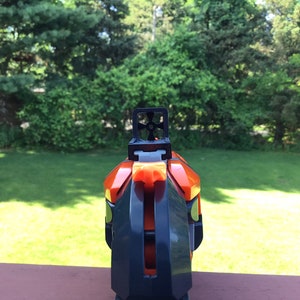 May include: A black and orange toy gun with a black sight. The gun is pointed towards a grassy area.