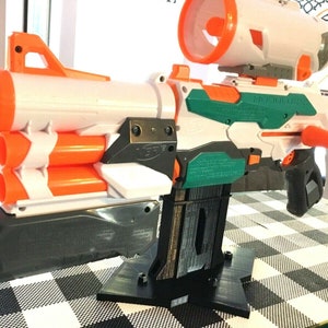 May include: A white and orange Nerf Modulus toy gun with a green barrel and a black stand. The gun has a scope on top and a magazine on the bottom.