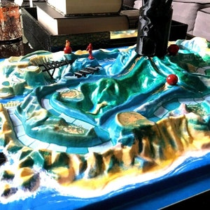 May include: A green and blue board game with a volcano in the center. The volcano has a black cone and red balls on the sides. There are small red and blue figures on the board.