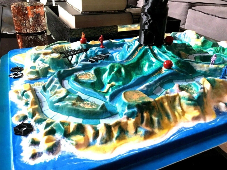 May include: A green and blue board game with a volcano in the center. The volcano has a black cone and red balls on the sides. There are small red and blue figures on the board.