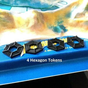 May include: Four black hexagon-shaped game tokens on a blue surface. The text "4 Hexagon Tokens" is printed below the tokens.