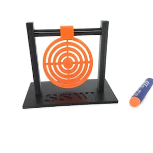 May include: A black target stand with an orange target and a blue dart. The target stand has a black base and two black vertical supports. The target is a circular orange disc with concentric rings. The dart is blue with a white tip and the word "Elite" printed on it.