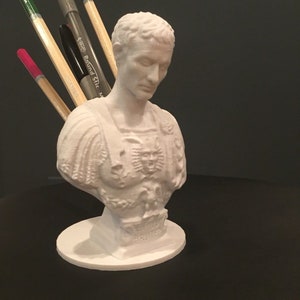 May include: White 3D printed bust of a Roman man with a pen holder in the chest area. The bust is on a round base and has the words "ET TU BRUTE" inscribed on the base.