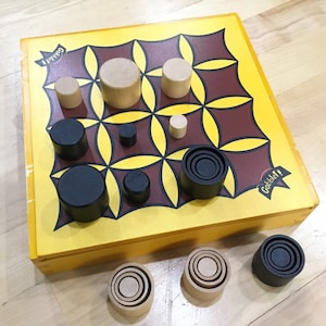 May include: A Gobblet Gobblers game board with black and brown pieces. The board is yellow with a brown and red pattern. The game is set up for play with pieces in various positions.