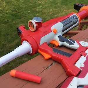 May include: A red and white toy gun with an orange trigger and a white barrel extension. A red and orange dart is lying on a wooden surface.