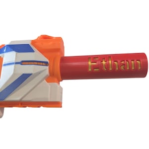 May include: A red and orange toy gun with a white and blue barrel. The barrel has the name "Ethan" engraved in gold letters.