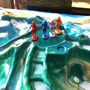 May include: Four plastic toy soldiers standing on a green and white game board. The soldiers are red, blue, orange, and purple.