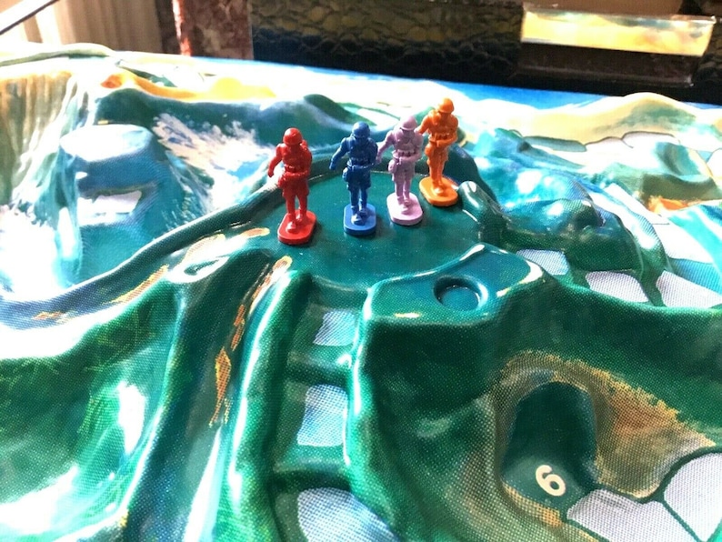 May include: Four plastic toy soldiers standing on a green and white game board. The soldiers are red, blue, orange, and purple.