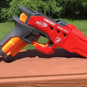 May include: A red, orange, and gray Nerf Mega BigShock blaster toy gun. The toy gun has the Nerf logo and the words "MEGA" and "BIGSHOCK" printed on the side. The blaster is made of plastic.