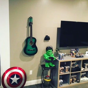 May include: A green acoustic guitar hangs on a wall next to a black television. A green Hulk figurine holds a video game controller. A shelf unit holds various figurines and video game related items. The shelf is labeled "Fortnite".