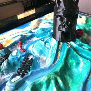 May include: A close-up of a board game with a black plastic skull-shaped piece in the center. The board is green and blue with red and orange game pieces.