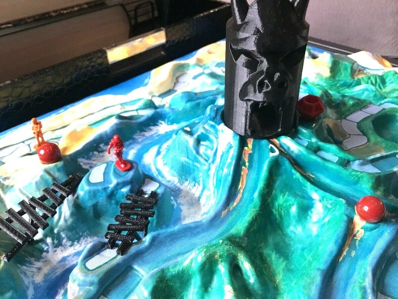 May include: A close-up of a board game with a black plastic skull-shaped piece in the center. The board is green and blue with red and orange game pieces.