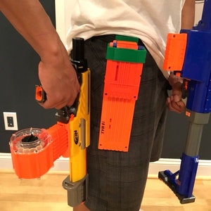 May include: A person wearing a gray and black plaid shorts with a blue and orange Nerf gun in their right hand. The gun has a clear orange magazine attached to the bottom. The person is also wearing a belt with multiple orange and green Nerf magazines attached to it. There is a blue and gray Nerf gun standing on the floor to the right of the person.