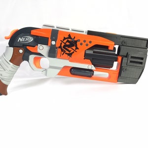 May include: A black and orange Nerf gun with a gray grip and a black barrel. The gun has a "NERF" logo on the side and a zombie-themed graphic on the top.