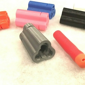 May include: A set of six 3D printed dart barrels in various colors, including orange, pink, blue, black, and gray. The barrels are designed with a three-pronged opening at the end. A red foam dart is also included.