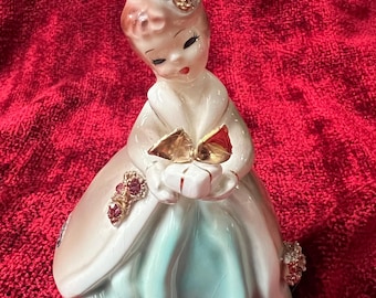 May include: A vintage porcelain figurine of a woman wearing a blue dress and a white coat, holding a gift box. She has a pink hat with rhinestones and a gold trim.