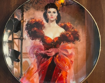 Peut inclure: Une assiette ronde en céramique avec un bord doré présente une peinture d'une femme vêtue d'une robe rouge et orange avec un col en plumes. La femme a les cheveux noirs et regarde directement le spectateur.