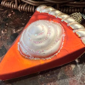 Peut inclure: Une d&eacute;coration de No&euml;l en verre en forme de part de tarte avec un tourbillon de gla&ccedil;age blanc sur le dessus. La cro&ucirc;te de la tarte est dor&eacute;e et la garniture est orange vif.