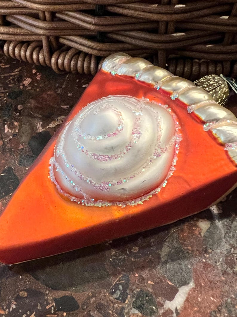 Peut inclure: Une d&eacute;coration de No&euml;l en verre en forme de part de tarte avec un tourbillon de gla&ccedil;age blanc sur le dessus. La cro&ucirc;te de la tarte est dor&eacute;e et la garniture est orange vif.