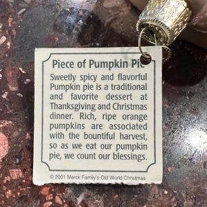 Peut inclure: Une &eacute;tiquette blanche avec du texte brun qui dit "Piece of Pumpkin Pie" et d&eacute;crit l'histoire de la tarte &agrave; la citrouille comme un dessert traditionnel de Thanksgiving et de No&euml;l. L'&eacute;tiquette est attach&eacute;e &agrave; une d&eacute;coration dor&eacute;e avec un motif d&eacute;coratif.
