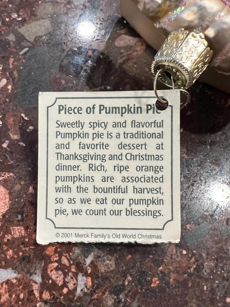 Peut inclure: Une &eacute;tiquette blanche avec du texte brun qui dit "Piece of Pumpkin Pie" et d&eacute;crit l'histoire de la tarte &agrave; la citrouille comme un dessert traditionnel de Thanksgiving et de No&euml;l. L'&eacute;tiquette est attach&eacute;e &agrave; une d&eacute;coration dor&eacute;e avec un motif d&eacute;coratif.