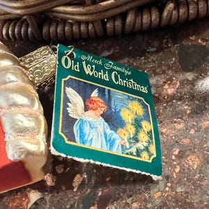 Peut inclure: Une &eacute;tiquette verte et blanche avec le texte "Merck Family's Old World Christmas" et une image d'un ange aux cheveux blonds et &agrave; la robe bleue debout devant un arbre avec des lumi&egrave;res blanches.