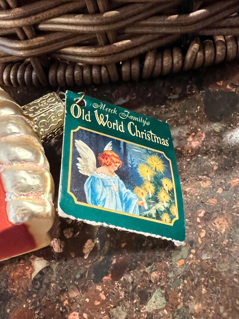Peut inclure: Une &eacute;tiquette verte et blanche avec le texte "Merck Family's Old World Christmas" et une image d'un ange aux cheveux blonds et &agrave; la robe bleue debout devant un arbre avec des lumi&egrave;res blanches.
