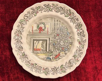 May include: A white ceramic Christmas plate with a green holly border. The center features a vintage illustration of a fireplace with a lit fire, a Christmas tree decorated with ornaments and presents, and a wreath hanging above the fireplace.
