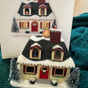 May include: Two miniature ceramic houses with snow-covered roofs, red doors, and lit windows. The houses are decorated with small trees and a star.