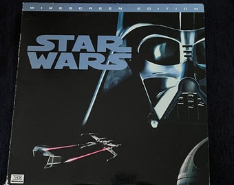 Peut inclure: Une jaquette de cassette VHS noir et blanc pour le film Star Wars. La jaquette présente le casque de Dark Vador et un chasseur TIE volant en arrière-plan. Le texte "WIDESCREEN EDITION" est en haut de la jaquette et le texte "STAR WARS" est au centre de la jaquette.