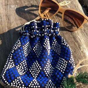 May include: A blue and white beaded drawstring bag with a fringe trim. The bag is made of a fabric with a geometric pattern. The bag is sitting on a wooden surface with a pair of brown sunglasses.
