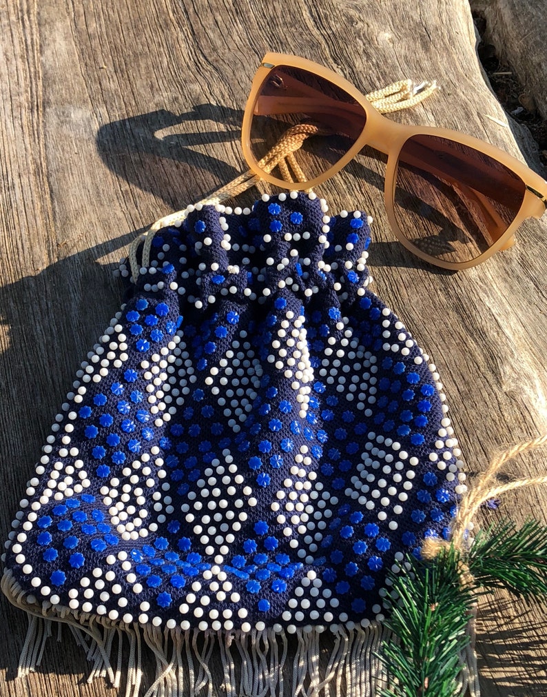 May include: A blue and white beaded drawstring bag with a fringe trim. The bag is made of a fabric with a geometric pattern. The bag is sitting on a wooden surface with a pair of brown sunglasses.