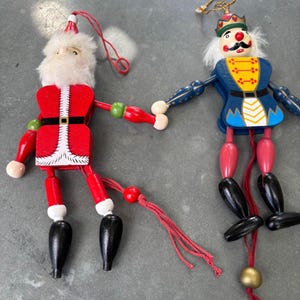 May include: Two wooden hanging ornaments featuring a Santa Claus figure in a red suit with white faux fur trim and a nutcracker soldier in a blue uniform with a crown. Both figures have articulated limbs controlled by red strings with beads.