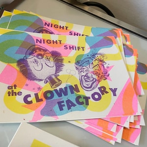 May include: A stack of square cards with a colorful clown-themed design. The cards feature the text "NIGHT SHIFT" and "at the CLOWN FACTORY" with illustrations of clown faces in bright pink, orange, yellow, and purple.