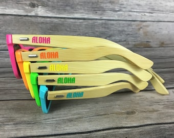 May include: A stack of sunglasses with bamboo-style frames and colorful accents. Each pair has a different color on the end of the frames, including pink, orange, yellow, green, and blue. The word "ALOHA" is printed on the side of each pair.
