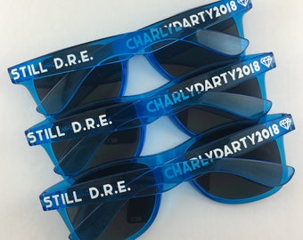 May include: Three pairs of blue plastic sunglasses with white text. The text on the sunglasses reads "STILL D.R.E." and "CHARLYDARTY2018".