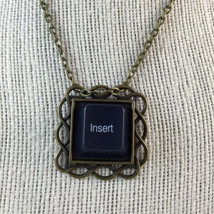 May include: A necklace with a bronze chain and a square pendant. The pendant features a black computer key with the word "Insert" on it.
