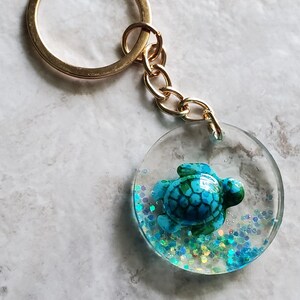 May include: A gold key chain with a round resin charm featuring a blue and green turtle with iridescent glitter inside.