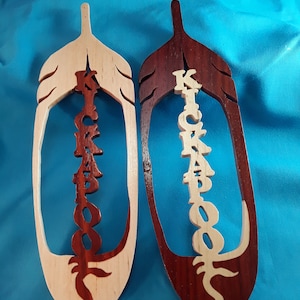 May include: Two wooden feather-shaped wall hangings. One is light-colored with dark brown lettering and accents, the other is dark brown with light-colored lettering and accents. The word "KICKAPOO" is carved vertically down the center of each feather.