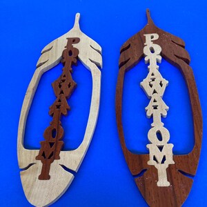 May include: Two wooden feather-shaped ornaments. One is light-colored with dark brown lettering, and the other is dark brown with light-colored lettering. The word "POTAWATOMI" is carved vertically down the center of each ornament.