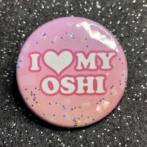 May include: A pink and white button with glitter. The button has a heart and the text "I love my Oshi" on it.