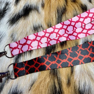 May include: Two patterned keychains with silver swivel clips. One keychain is pink with a red geometric pattern. The other keychain is black with a red geometric pattern.