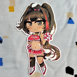 Puede incluir: Una pegatina troquelada con una animadora de dibujos animados con cabello castaño y reflejos rojos. Lleva un uniforme de animadora rojo y blanco con la palabra "CHEER" en la parte superior. La pegatina incluye un pompón y está sobre un fondo estampado.