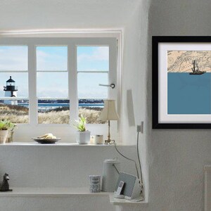 May include: A framed art print featuring a nautical theme. The artwork depicts a sailboat on a blue sea, set against a map background. The frame is black, and the print is displayed on a white wall. A window with a lighthouse view is also visible.
