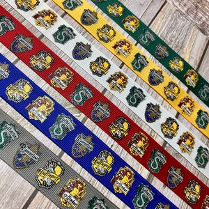 Op de afbeelding: Assortiment linten met verschillende huisschilden uit een fantasieserie. De linten zijn in meerdere kleuren, waaronder groen, geel, rood, blauw, wit en grijs. Elk lint toont een ander huisschild met de huisnaam.