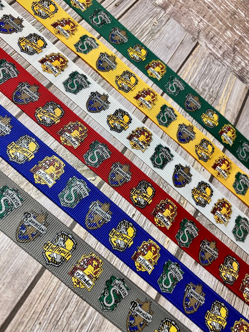 Op de afbeelding: Assortiment linten met verschillende huisschilden uit een fantasieserie. De linten zijn in meerdere kleuren, waaronder groen, geel, rood, blauw, wit en grijs. Elk lint toont een ander huisschild met de huisnaam.