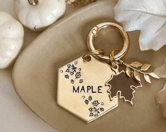 May include: Gold-toned keychain with a hexagonal charm featuring the word "MAPLE" and a small maple leaf design. The charm is attached to a gold-toned key ring with a smaller gold-toned charm of a maple leaf with a brown enamel finish.