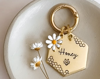 May include: Gold hexagon-shaped keychain with a bee and honeycomb design. The keychain has the word "Honey" engraved on it and is adorned with a white daisy with a yellow center.