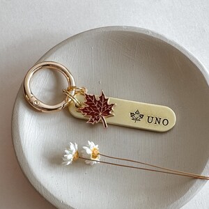 May include: A gold-colored keychain with a maple leaf charm and the word "UNO". The maple leaf is red and the keychain is attached to a gold ring. The keychain is resting on a gray, circular dish.